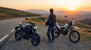 Man with helmet stands between a blue roadster and a white trail bike on a winding mountain road at golden hour, scenic backdrop.