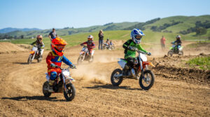 Young children in colorful motocross gear ride dirt bikes on a sunny track. A toddler on a balance bike leads, older child kicks up dust.